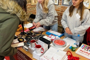 Dwie kobiety obsługują stoisko z materiałami promocyjnymi i gadżetami, rozmawiając z osobą stojącą po drugiej stronie stołu. W tle widoczne są obrazy na ścianie z cegły.