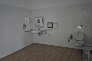 Pomieszczenie z białymi ścianami i podłogą w kolorze drewna. Na ścianie widoczne przyłącza instalacyjne i elementy montażowe. Brak mebli. Pomieszczenie przygotowane pod zabudowę kuchenną lub techniczną.