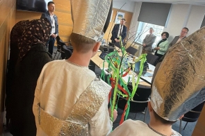 Dzieci w strojach ludowych stoją tyłem, trzymając gałązkę z kolorowymi wstążkami, naprzeciw nich stoją dorośli w pomieszczeniu z stołem i krzesłami.