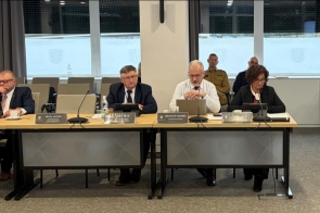 Osoby siedzą przy stole z mikrofonami, dokumentami i tabletami, w tle widoczne są okna z zasłonami oraz siedzące osoby.