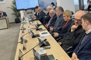 Osoby siedzą przy stole z mikrofonami, tabletami, dokumentami i filiżankami, patrząc w stronę ekranu.