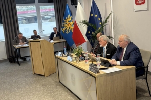 Kilku mężczyzn siedzi przy stołach z mikrofonami i dokumentami, w tle stoją flagi Polski, Unii Europejskiej oraz Powiatu Cieszyńskiego.