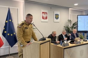 Mężczyzna w mundurze służbowym stoi przy mównicy i czyta dokument, obok znajduje się stół z mikrofonami i dokumentami, a w tle widoczne są flagi oraz herby na ścianie.