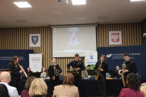 Przed siedzącymi na krzesłach, uczestnikami konferencji, stoi pięciu muzyków ubranych na czarno, w rękach trzymają instrumenty.