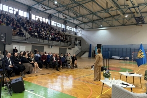 Kobieta stoi na środku sali gimnastycznej i przemawia do siedzącej publiczności, a na trybunach widać liczne osoby oglądające wydarzenie.