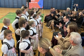Dzieci w strojach ludowych stoją w rzędzie naprzeciw siedzącej publiczności, która bije brawo na sali gimnastycznej.
