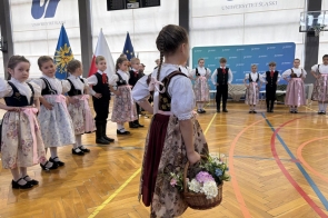 Dzieci w strojach ludowych stoją w kole na sali gimnastycznej, a jedna dziewczynka z koszykiem kwiatów stoi na pierwszym planie.