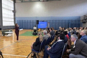 Kobieta stojąca na sali gimnastycznej przemawia do siedzącej publiczności, a za nią widać ekran i osobę przy stole.
