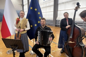 Trzy osoby w strojach ludowych stoją na sali gimnastycznej z instrumentami – skrzypcami, akordeonem i kontrabasem – na tle flag.