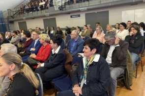 Publiczność siedzi na krzesłach na sali gimnastycznej i słucha wydarzenia, a w tle widać kolejne rzędy osób oraz trybuny.