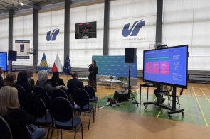 Kobieta przemawia do siedzącej publiczności na sali gimnastycznej, obok widać ekran z prezentacją oraz ustawione flagi.