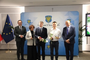 Pięciu mężczyzn i jedna kobieta stoją do zdjęcia na tle ścianki powiatu, dwaj z nich trzymają kwiaty i upominki.