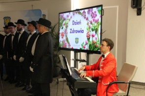 Mężczyzna w czerwonej marynarce gra na pianinie, obok stoi grupa elegancko ubranych śpiewaków.
