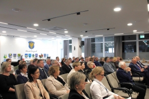 Publiczność siedzi w sali konferencyjnej i uważnie słucha wydarzenia, na ścianie widoczny jest herb Powiatu Cieszyńskiego i rzędy herbów gmin.