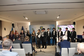 Grupa osób w eleganckich, stylizowanych strojach stoi na scenie, część z nich ma kapelusze i rękawiczki. 