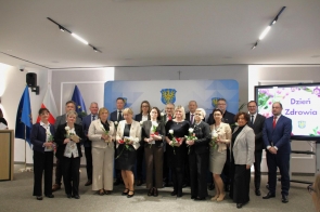 Grupa kilkunastu osób stoi do wspólnego zdjęcia na tle ścianki powiatu, w centrum jedna z kobiet trzyma bukiet kwiatów.