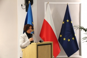 Janina Żagan Starosta Cieszyński stoi przy mównicy i przemawia do publiczności, trzymając mikrofon, a za nią widać flagi