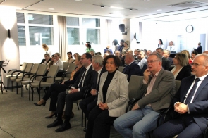 Grupa elegancko ubranych osób siedzi w sali konferencyjnej i z uwagą słucha wydarzenia, część patrzy w stronę sceny.
