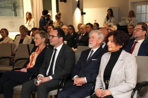 Rząd elegancko ubranych osób siedzi na krzesłach i uważnie słucha wydarzenia, a za nimi widać większą publiczność.