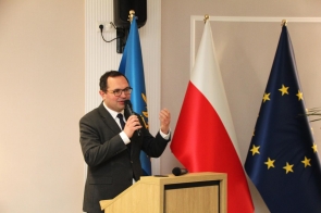 Michał Kopański Wojewoda Śląski w garniturze przemawia przy mównicy, trzymając mikrofon, a za nim stoją flagi.