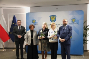 Pięć osób stoi do wspólnego zdjęcia na tle ścianki z herbem powiatu, dwie kobiety trzymają kwiaty i upominki.