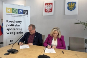 Przy stole siedzą kobieta i mężczyzna, którzy podpisują dokumenty. W tle widoczny jest baner Regionalnego Ośrodka Polityki Społecznej.