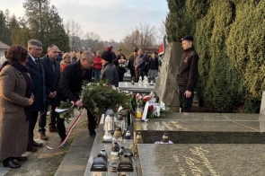 Mężczyzna składa wieniec przy grobie ozdobionym zniczami i kwiatami. Obok stoją pozostali uczestnicy.