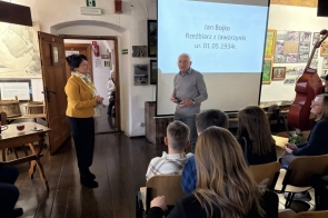 Jan Bojko stoi przed zebranymi i przemawia, a za nim widoczny jest ekran. Uczestnicy siedzą na krzesłach i słuchają, a obok stoi kobieta.