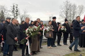 Grupa osób stoi na cmentarzu, trzymając wieńce i wiązanki kwiatów ozdobione biało-czerwonymi wstęgami.
