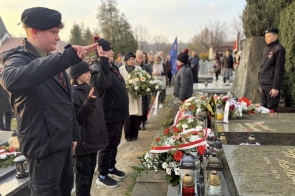 Młodzi chłopcy stoją przy grobie udekorowanym kwiatami, wieńcami i zniczami oraz wykonują gest salutowania.