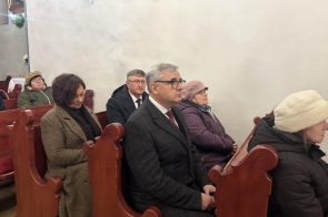 Uczestnicy siedzą w ławkach kościoła. Widać kilka osób, ubranych w płaszcze, zwróconych w stronę ołtarza.