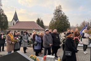 Grupa osób stoi na cmentarzu, trzymając wieńce i wiązanki kwiatów.