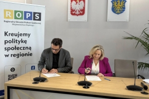 Przy stole siedzą kobieta i mężczyzna podpisujący dokumenty. Obok znajduje się baner.