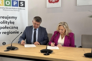 Przy stole siedzą kobieta i mężczyzna podpisujący dokumenty. Obok znajduje się baner.
