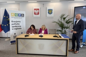 Przy stole siedzą dwie kobiety podpisujące dokumenty, obok stoi mężczyzna trzymający teczkę.