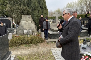 Mężczyzna stojący przy grobie trzyma mikrofon i przemawia. Wokół znajdują się nagrobki, znicze i kwiaty.