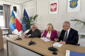 Przy stole siedzą trzy osoby, które podpisują dokumenty. W tle widoczne są flagi.