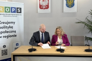 Przy stole siedzą kobieta i mężczyzna podpisujący dokumenty. Obok znajduje się baner.
