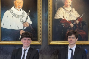 Dwóch młodych mężczyzn ubranych elegancko w garnitury, siedzących przy stole. Za nimi wiszą dwa duże, klasyczne portrety przedstawiające starszych mężczyzn w uroczystych strojach akademickich.