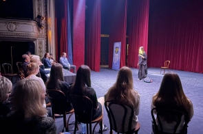 Osoba stojąca na scenie teatru i przemawiająca do siedzącej publiczności, w otoczeniu czerwonych kurtyn.
