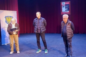Trzy osoby stojące na scenie teatru, jedna trzyma dokumenty, w tle czerwona kurtyna, plakat oraz baner.