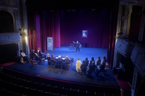Widok sceny teatralnej z perspektywy widowni: jedna osoba występuje na środku, przed nią siedzi grupa osób przy stoliku i na krzesłach.