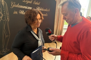 Janina Żagan Starosta Cieszyński trzyma dokument i rozmawia z mężczyzną z mikrofonem. W tle czarna ścianka z napisem o zespole szkół gastronomiczno-hotelarskich.