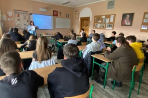 Zdjęcie przedstawia salę lekcyjną z uczniami siedzącymi w ławkach. Uczniowie zwróceni są w stronę tablicy, gdzie wyświetlana jest prezentacja. Na ścianach widoczne są plakaty edukacyjne. W klasie znajdują się ławki, krzesła i sprzęt szkolny.