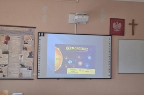 Zdjęcie przedstawia salę lekcyjną. Na ścianie wyświetlony jest slajd z prezentacji „Dermokosmos”. Przed ścianą ustawione są ławki i krzesła. Po lewej stronie widoczne plakaty edukacyjne, po prawej tablica.