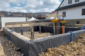 Wykop z fundamentami i wystającym zbrojeniem, obok budynek i niewielka maszyna budowlana, w tle zabudowa i wzgórza.