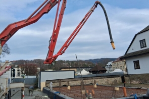 Ramię pompy do betonu nad placem budowy, przygotowane zbrojenie fundamentu, obok zabudowania i pagórkowaty krajobraz.