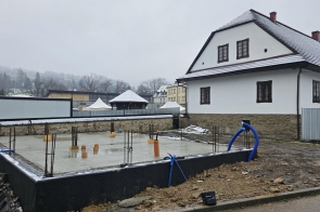 Plac budowy z wylaną płytą fundamentową i wystającymi prętami, obok biały budynek, w tle zabudowa i ośnieżone wzgórze.
