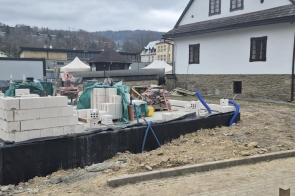 Plac budowy z wznoszonymi ścianami z białych bloczków, rozłożone materiały i sprzęt, obok budynek i zabudowa w tle.
