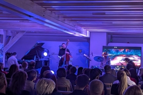 W sali siedzi publiczność, a na scenie kilku muzyków gra na instrumentach, w tle widoczny jest ekran z wizualizacją.